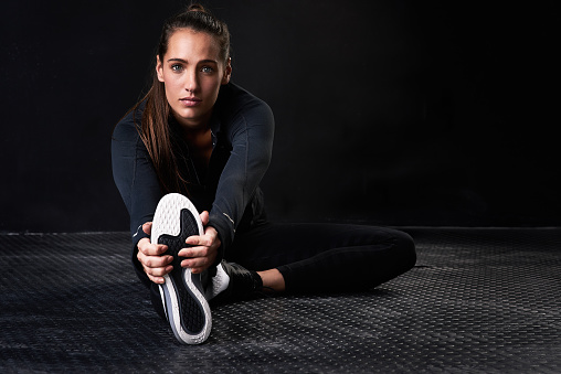 Studio portrait of a young woman in gym clothes stretching her legs against a dark background