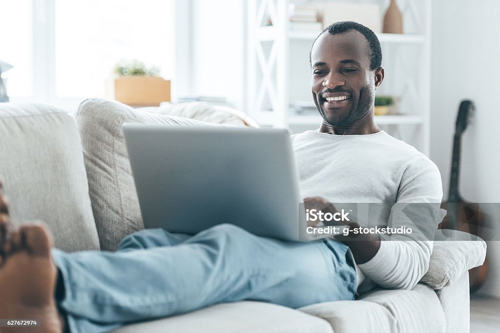 Relaxation at home. Handsome young African man looking at laptop and smiling while lying on the sofa at home Laptop Stock Photo