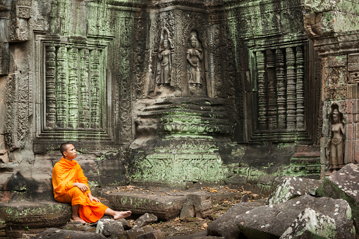 Contemplative Cambodian monk in an orange robe sits on the ancient steps of a temple ruin lost in thought looking at ancient carvings, Siem Reap, Cambodia, Asia