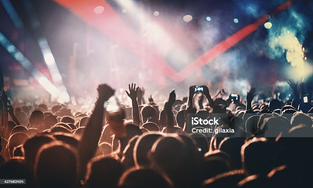 Rock concert. Rear view of people cheering at concert during music festival.Large group of unrecognizable adults with their arms raised.Some of them holding beer cans,some are taping the show with phones.There's a stage and a band performing in background,out of focus.Stage is toned purple blue with lasers and spotlights cutting through smoke and dust. Music Festival Stock Photo