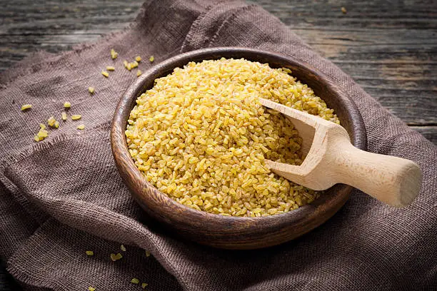 Raw bulgur in wooden bowl on brown textile, close up shot. Healthy, dietary, vegan, gluten free product