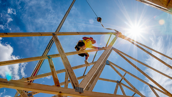 Manual laborer poised on a new construction hammering a piece of frame work with The sun and a partly cloudy blue sky in the background.