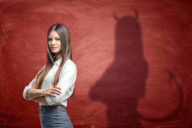 Young businesswoman is casting shadow of devil on rusty orange Young businesswoman is casting shadow of devil on rusty orange wall behind her. Unfair competition. Aggressive marketing strategies. Hidden motives. horned photos stock pictures, royalty-free photos & images