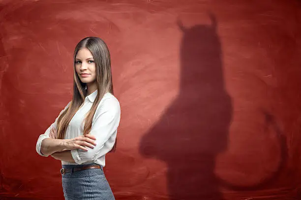 Young businesswoman is casting shadow of devil on rusty orange wall behind her. Unfair competition. Aggressive marketing strategies. Hidden motives.