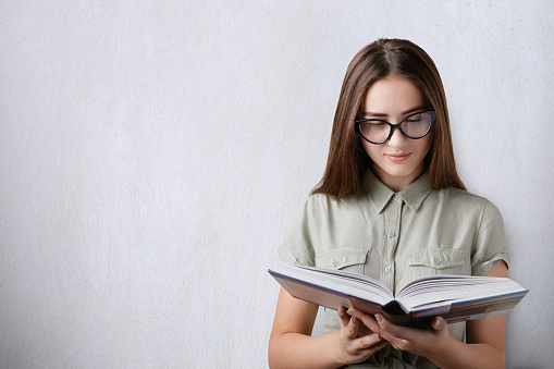 Education and studying. A portrait of a clever beautiful girl with long dark hair wearing glasses holding and reading a hardcover book on white background. An attentive reading of the book