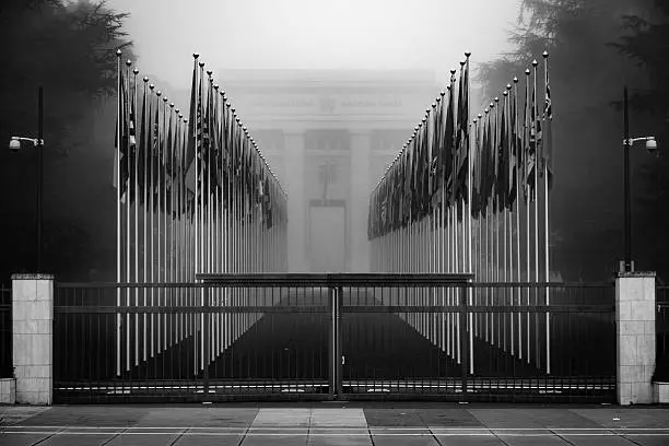 This is a black and white photo of the entrance to the United Nations in Geneva Switzerland; the gate is closed and beyond the gate are the flags blanketed by a dense fog. 