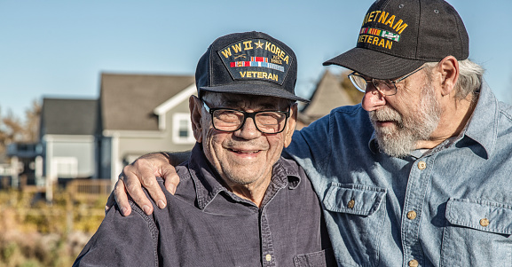 Two generations of real person senior adult men USA military veterans. On the left, a 93 year old United States Army World War II and Korean Conflict US Air Force military veteran is smiling at the camera while his son-in-law on the right - a 65 year old US Navy sailor Vietnam War military veteran - is teasing him - trying to distract him by telling jokes during the photo shoot. They are both wearing generic souvenir shop military veteran commemorative baseball style caps.