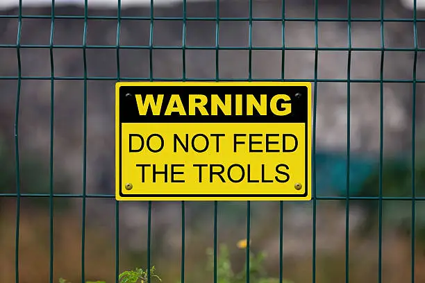Black and Yellow warning sign attached on a fence. The sign stating “Warning - Do not feed the trolls”.