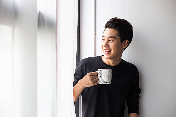 jeune homme asiatique tenant tasse - homme mug regarder dehors photos et images de collection