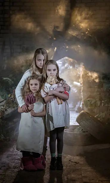 Three cute sad little girls of different ages dressed in a long white shirts. The older girl hugging little girls. The middle age girl holding a doll in crossed hands. The younger girl holding a big flower. The children are sad looking at the camera. The girls standing on background of a shack from old boards and rubbish. Ideas on homeless orphans in post apocalyptic world. Shooting at the old abandoned building in the slum