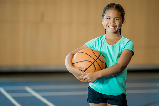 An elementary age girl is playing basketball at the gym. She is smiling and looking at the camera.