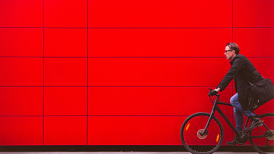 Handsome man, on the way to work, riding bicycle beside the red wall. The man is casually dressed and wears eyeglasses and carries black briefcase hung on shoulder. Motion concept, motion blur, copy space has been left.