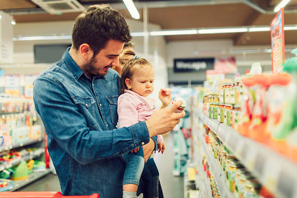 Father And His Baby Daughter Groceries Shopping Father and his baby girl groceries shopping. He is carrying his daughter and reading nutrition label on baby food product. baby food stock pictures, royalty-free photos & images