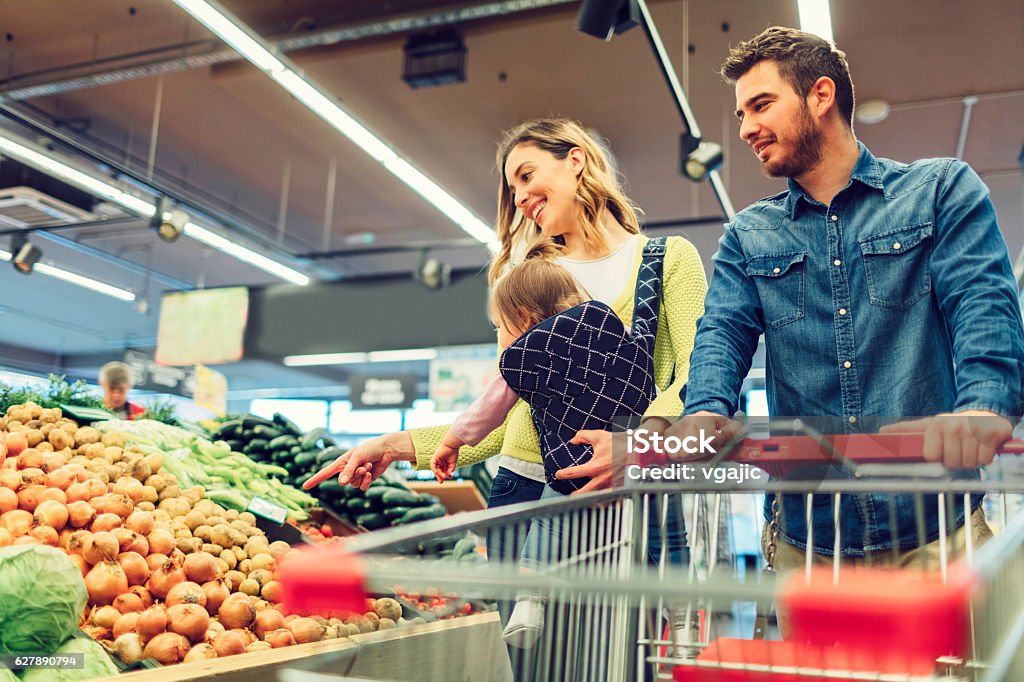 Family In Local Supermarket. Young family with one child in the shopping. Mother carry their baby daughter in baby carrier. They are in supermarket buying groceries. Choosing vegetables. Retail Stock Photo