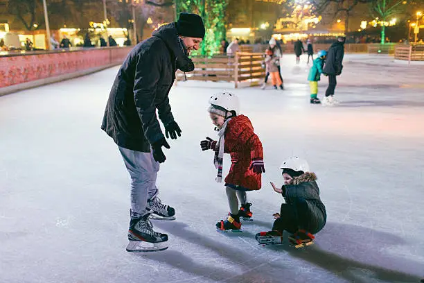 Photo of a little boy and his sister learning how to ice-skate on Ice rink. While their father is showing them basic positions, little boy fell down.