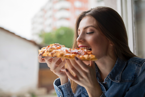 Beautiful, young woman leaning at the window, eating pizza and enjoying