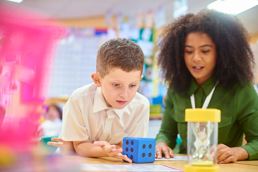 a primary aged schoolboy sits with his teacher in a one to one maths session . He is tossing a big dice used to add fun to the maths that he is struggling with  .Also one the desk is an hourglass to aid the visual appeal and stimulation of the task for the student.