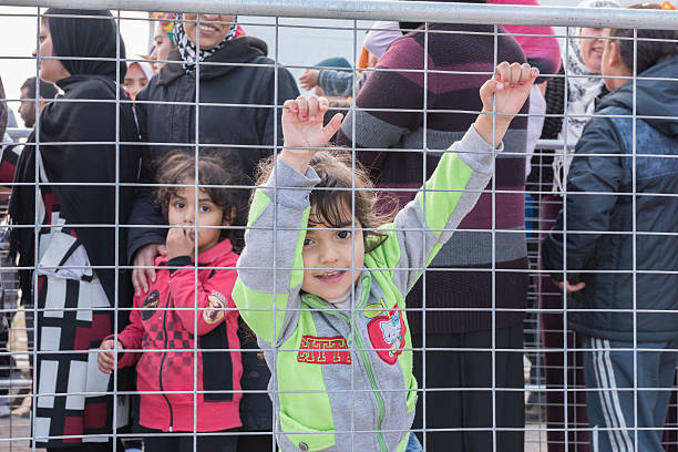Chios, Greece - May 5, 2016: Refugees are waiting in line for being served breakfast in Souda refugee camp in Chios Town on Chios Island in Greece. There are two separate lines; one for women and children and one for men. Pictured is the line for women and children. In the foreground is a little girl climbing the fence. In the background is a line of refugees that include the girl's mother and sister. The refugees have landed on Chios after crossing the Aegean Sea in flimsy inflatable boats from the shore of Turkey. As Europe and EU close their borders, the immigrants are stuck in Greece. The meals are financed by the Norwegian NGO Drop in the Ocean and served by its volunteers. At the moment 1,250 refugees are fed each day.