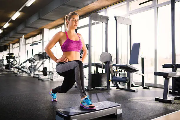 Photo of Woman doing lunges on step aerobics equipment at gym.