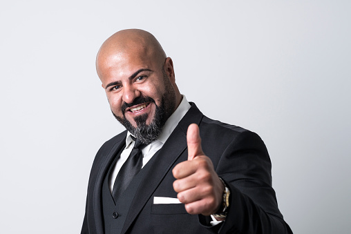 Bald businessman with black beard looking happy at camera with his thumb up. He is wearing an elegant black suit, a white button-down shirt and a tie.