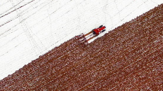Aerial view of tractor tilling snow covered field in Winter.