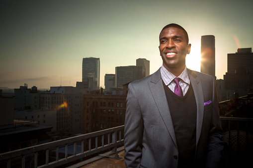 Grinning young black man in a suit on a downtown balcony.