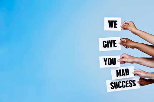 Five hands hold up words, vertically stacked,  saying 'We give you mad success' against a sky-blue background with ample copy space.
