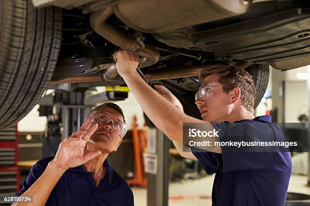 Mechanic And Male Trainee Working Underneath Car Together Stock Photo - Download Image Now