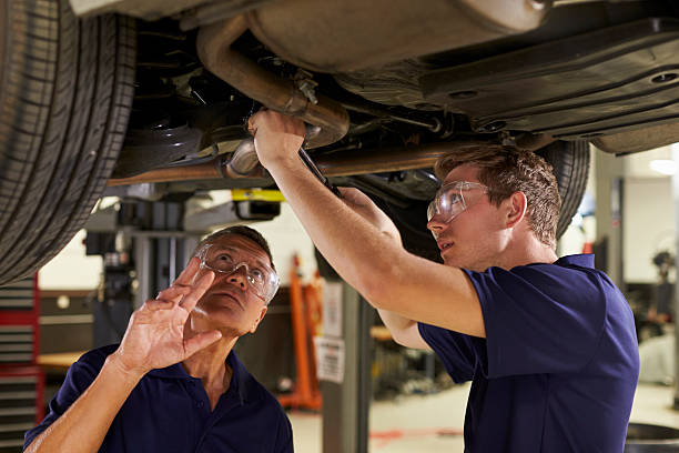 mecânico e estagiário masculino trabalhando debaixo do carro juntos - auto repair shop mechanic garage workshop - fotografias e filmes do acervo