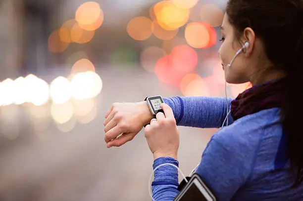 Beautiful young woman in the city with smart phone, smartwatch and earphones, listening music. Using a fitness app for tracking weight loss progress, running goal or summary of her run. Rear view.