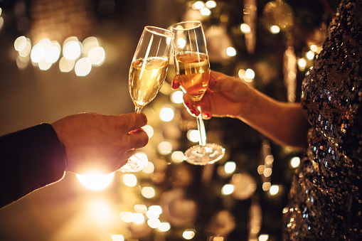 Couple holding glasses of champagne in front of Christmas tree. Toasting to each other. Evening or night.