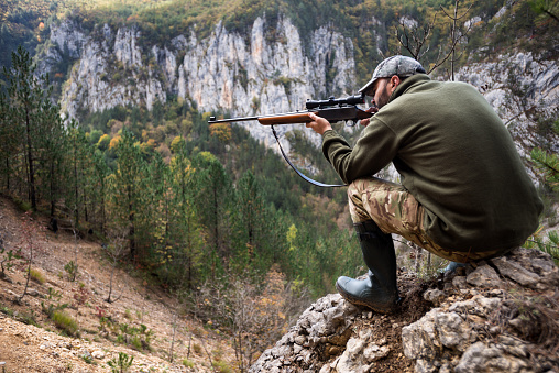 Portrait of a hunter crouching on top of the hill and aiming with rifle.