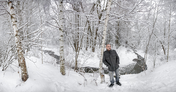 Mature man with shaved head dressed in casual clothing. The man is leaning his shoulder to birch. He is standing with his hands in his jacket pockets. The man pensively looking away. Shot in the woods on a winter day, wide angle shooting