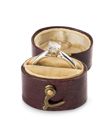A sparkling solitaire diamond ring is in vintage red leather ring box on a velvet cushion. The 1-carat diamond is set on a platinum band. The ring box is on a white background with a clipping path.