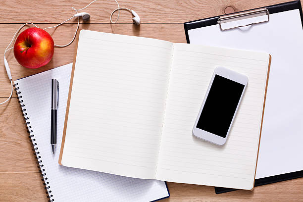Notepad, diary, mobile phone and apple fruit. Office or student's devices on modern wooden desk. Working table top view. Education or job background with copy space on paper sheet