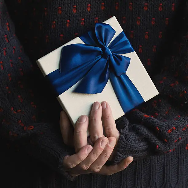 Close up of a man in knitted dark sweater holding a white present gift box with blue ribbon behind his back