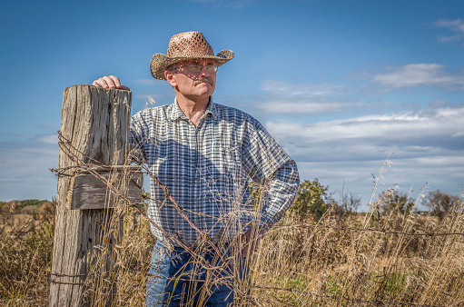 horizontal image of a caucasian white male farmer wearing a cowboy hat standing and leaning against a fence post gazing into the distance on a warm fall day.