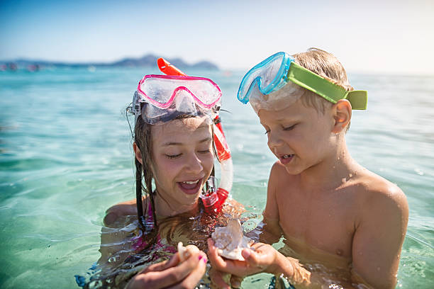 Little boy and his elder sister having fun snorkeling in beautiful sea. The kids aged 6 and 10 are examining sea shells found in sea. Sunny summer day.