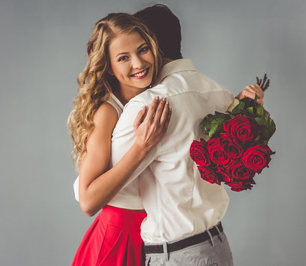 Beautiful elegant couple is hugging and smiling, on gray background. Girl is holding roses