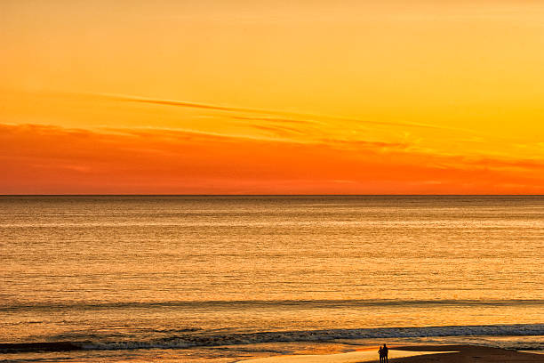 Beach and ocean image with unrecognizable people silouetted against the ocean.  Sunset sky colors are reflected in ocean waves.