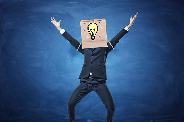 Businessman standing with cardboard box on his head Businessman standing with cardboard box on his head with a drawn electric bulb on blue blackboard background. Brainstorming. Business development. Solving problems. Anonymous person. man with a light bulb over his head stock pictures, royalty-free photos & images
