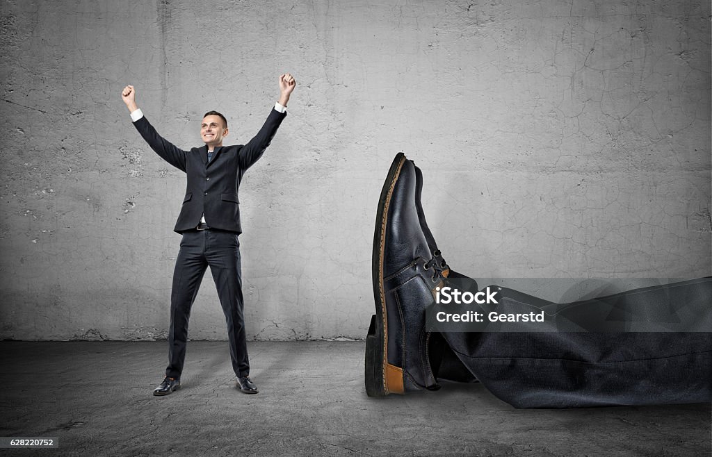 Small businessman standing with his arms up near giant leg A small businessman standing with his arms up and smiling happily near a giant leg in trousers and shoe lying near him, on the concrete gray background. Winning contest. Best in field. David and Goliath. Small company wins big one. Large Stock Photo