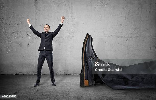 Small Businessman Standing With His Arms Up Near Giant Leg Stock Photo - Download Image Now