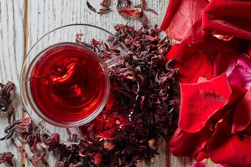 Red Hot Hibiscus tea in a glass mug on a wooden table among rose petals and dry tea custard with metallic heart