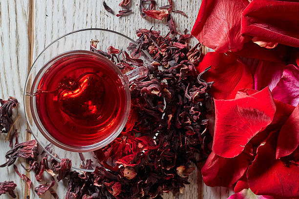 Red Hibiscus tea in glass mug Red Hot Hibiscus tea in a glass mug on a wooden table among rose petals and dry tea custard with metallic heart pink tea stock pictures, royalty-free photos & images
