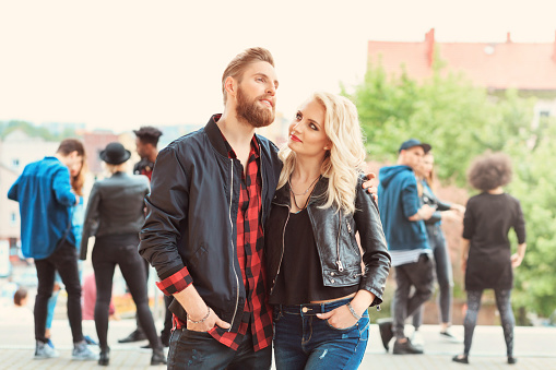 Bearded young man and beautiful blonde young woman embracing outdoor. Group of people in the background.