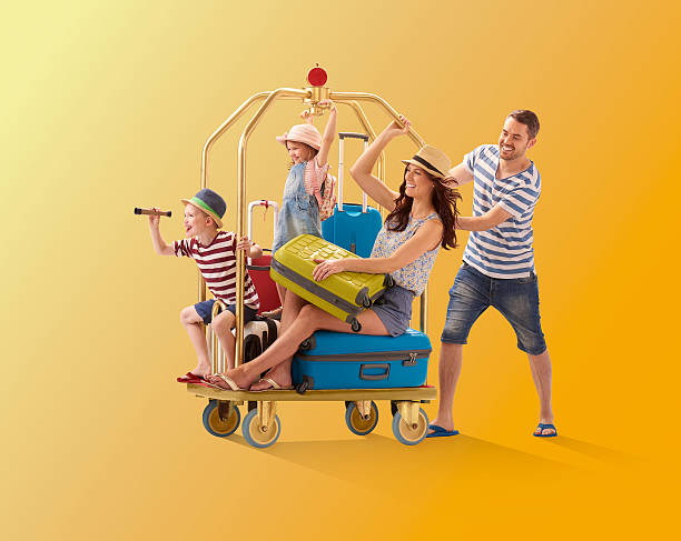 off on holiday a young family climb aboard a luggage trolley and dad pushes them off to their holiday destination . They are all wearing warm weather clothing and holding their suitcases. bellhop photos stock pictures, royalty-free photos & images