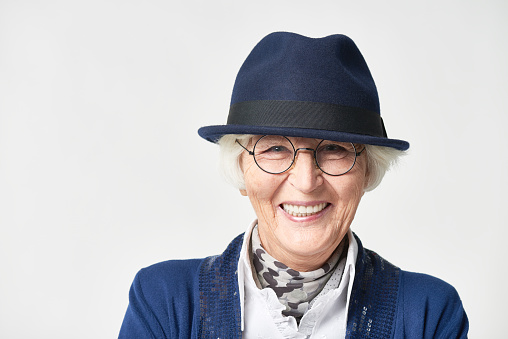 Close-up portrait of fashionable elderly woman in round glasses and hat looking at camera and smiling brightly, showing beautiful teeth on white wall background