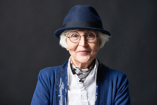 Close-up portrait of stylish old lady in round glasses and hat standing on dark wall background, looking at camera confidently and smiling slightly