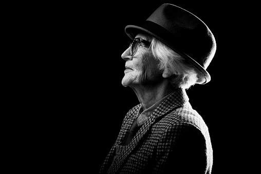 Profile view of wrinkled senior woman in fedora hat and trendy eyeglasses looking away against black background, black and white portrait shot